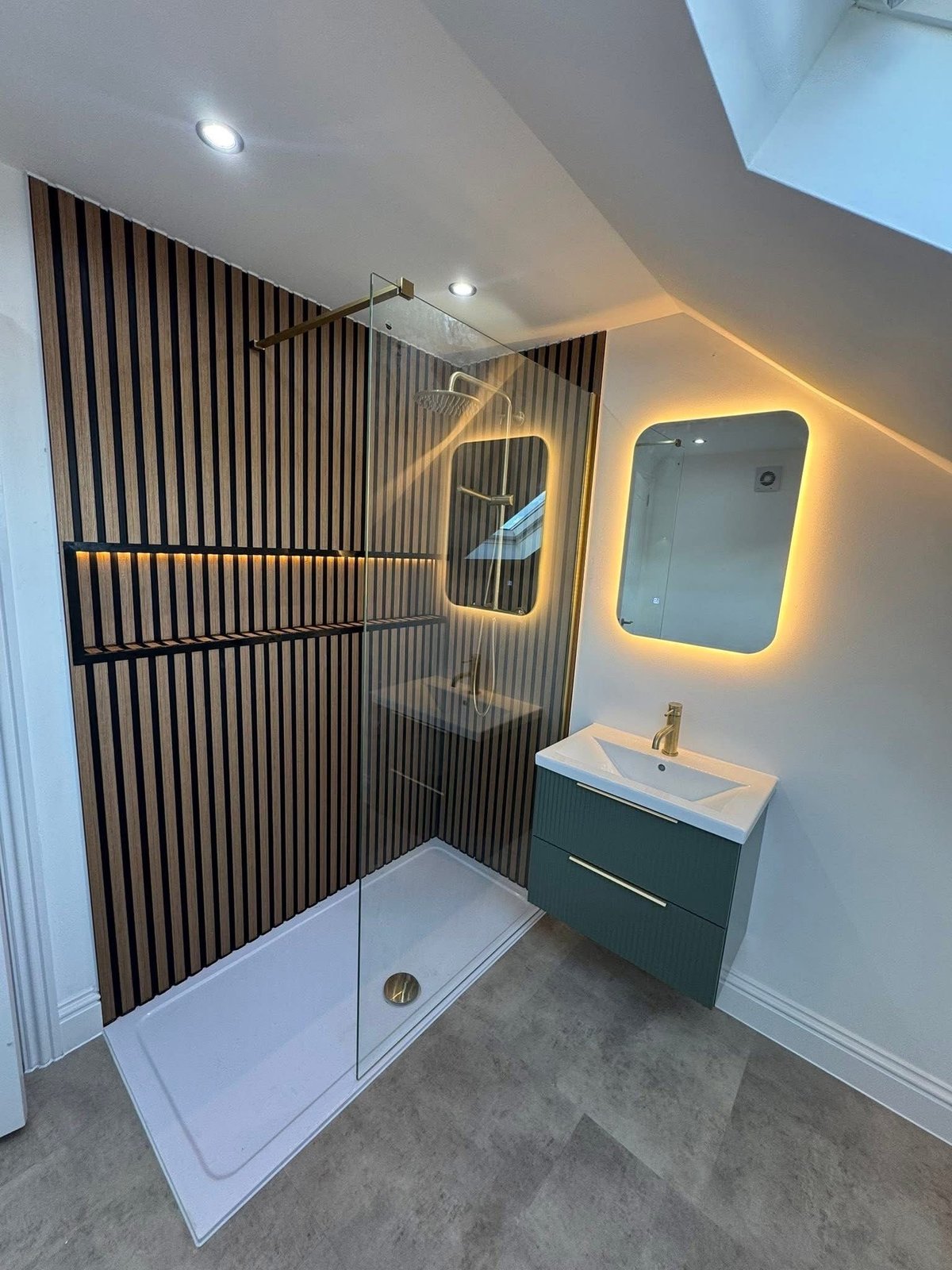 Modern bathroom with walk-in shower, black vertical tile wall, illuminated mirrors, green vanity with white sink, and gray flooring