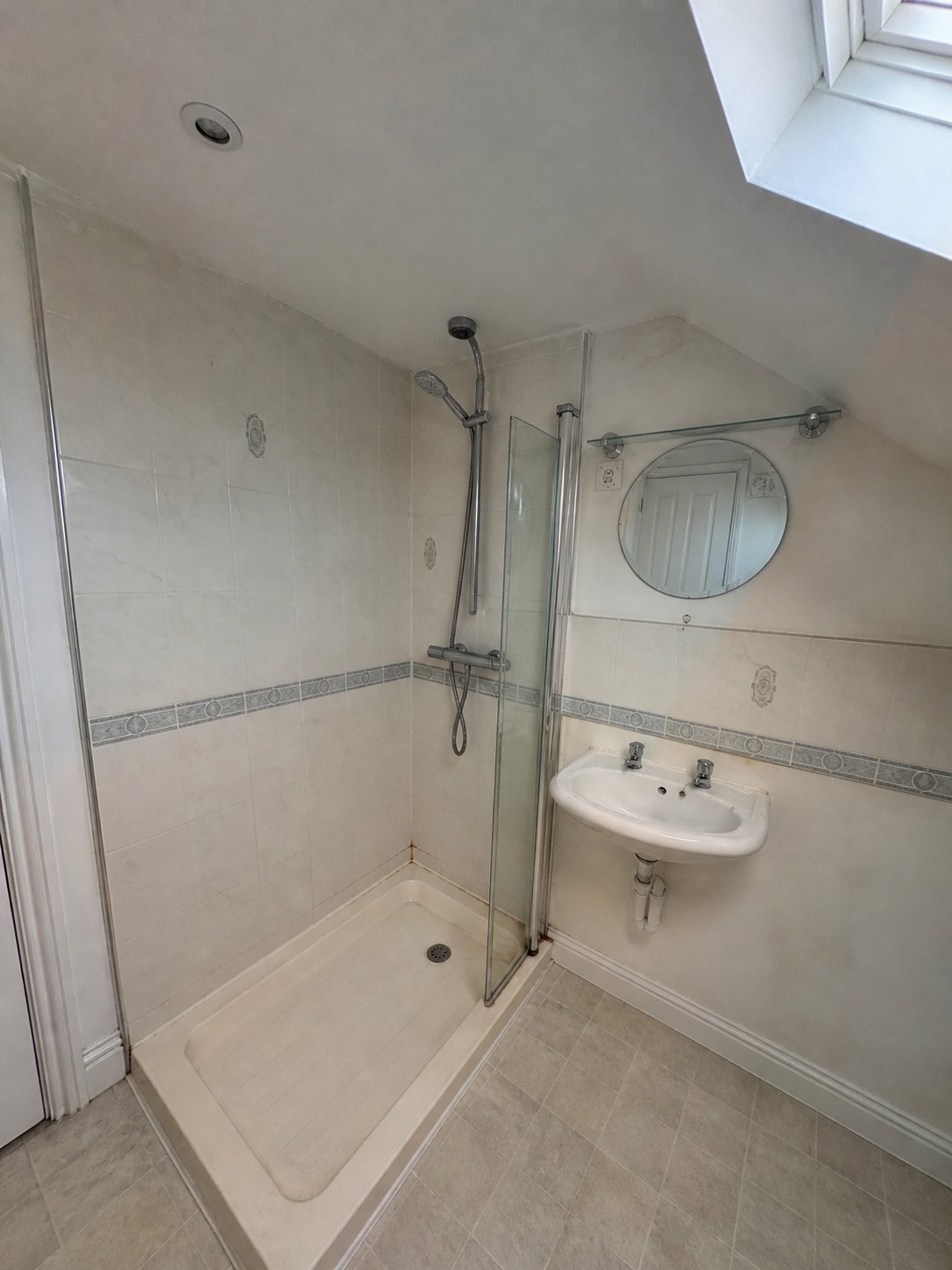 Modern bathroom with glass-enclosed shower, white wall tiles, circular mirror, and wall-mounted sink under skylight